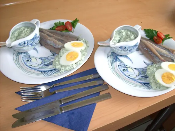 Zwei Teller mit frischer Matjes, gehackten Eiern und Grie Soß auf einem Holztisch, umgeben von Besteck und frischen Tomaten.