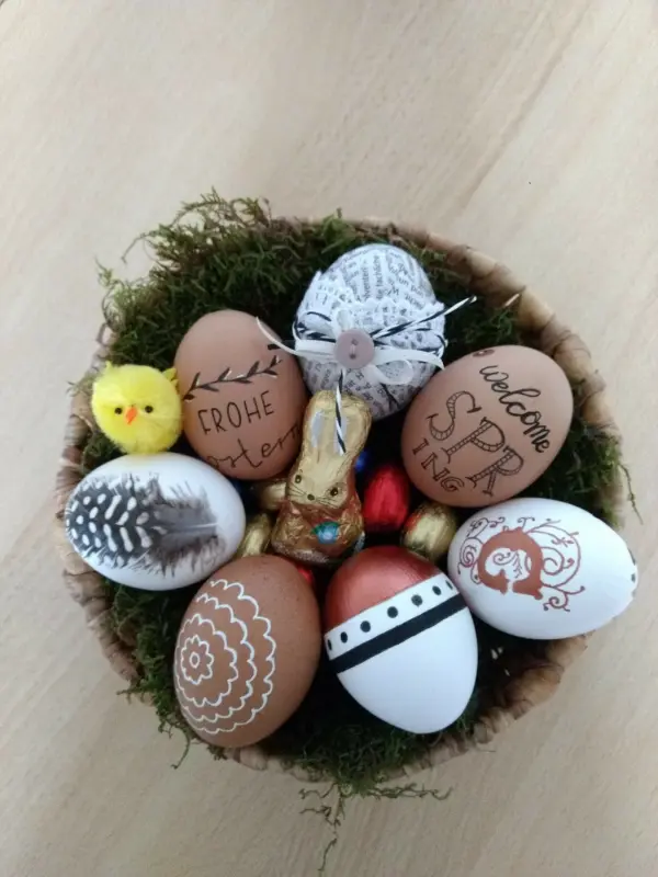 In einem Korb liegen kunstvoll verzierte Eier mit verschiedenen Mustern und einem kleinen Küken, umgeben von grünem Moos.