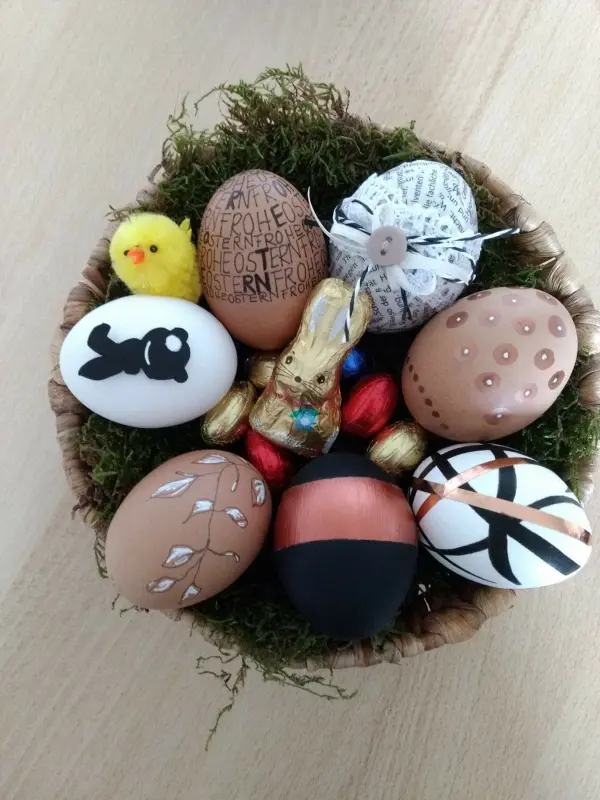 Bunte, detailreich verzierte Eier liegen in einem Korb auf Moos, umgeben von kleinen Schokoladenhasen und gelben Küken.
