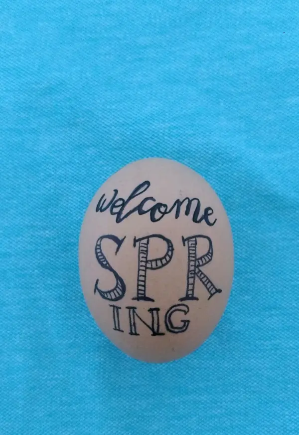 Ein dekoriertes Ei liegt auf einem hellblauen Stoff und trägt die Aufschrift „welcome SPRING“. Es symbolisiert vorösterliche Freude.