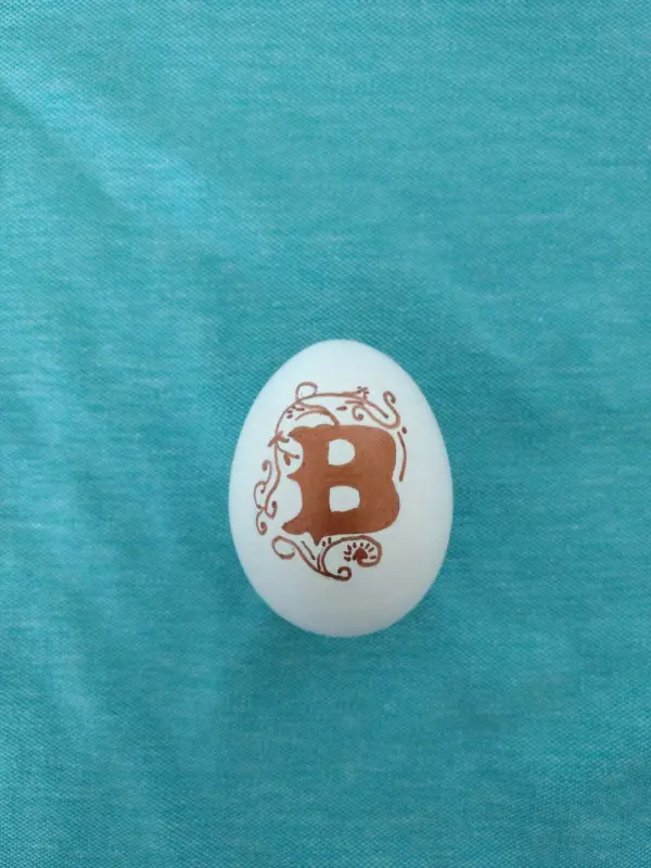 Ein weißes Ei mit einer braunen, kunstvollen "B"-Verzierung liegt auf einem hellblauen Stoffhintergrund.