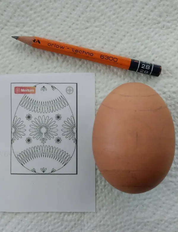 Ein Ei liegt neben einer Vorlage mit einem detaillierten Muster und einem Bleistift; es wird für die Verzierung vorbereitet.