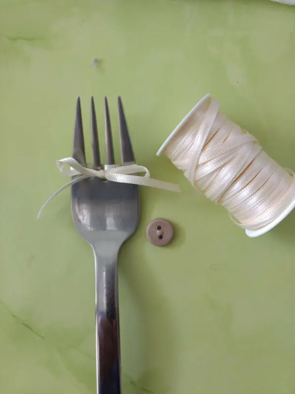 Eine Gabel liegt mit einem kleinen weißen Schleifenband darauf, daneben eine Spule mit glänzendem Faden und einem Knopf auf einem grünen Untergrund.