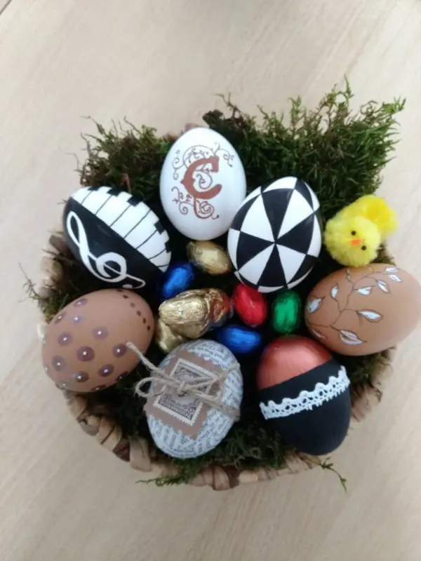 Bunte Eier liegen in Moos, kunstvoll verziert, umgeben von kleinen Schokoladen-Eiern und einem gelben Küken.