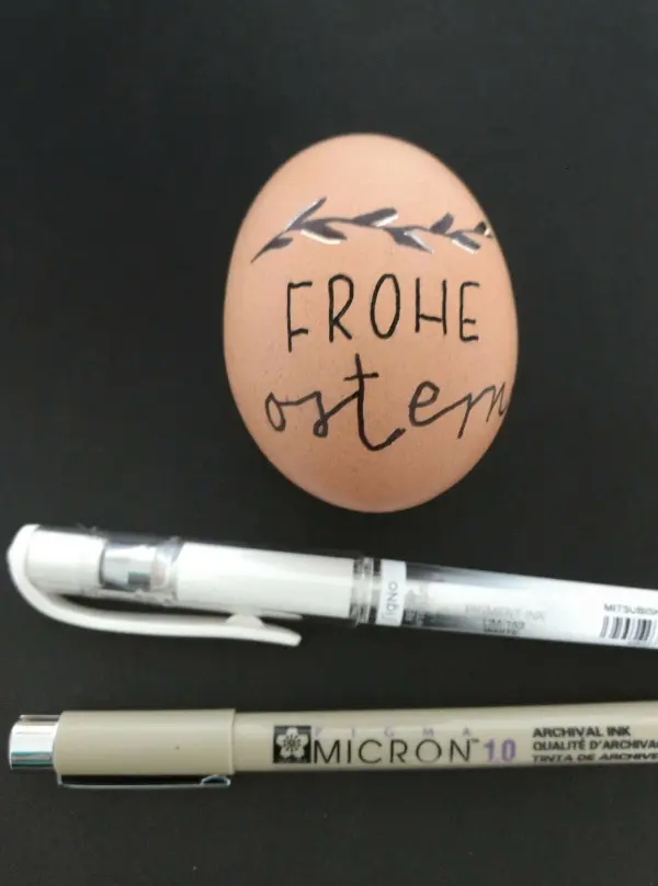 Ein dekoriertes Ei liegt auf einem schwarzen Untergrund, beschriftet mit „FROHE OSTERN“, umgeben von Malstiften.
