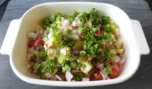 Ein Wurstsalat in einer weißen Schüssel wird mit frischem Gemüse wie Radieschen und Salat zubereitet und steht auf einem dunklen Untergrund.