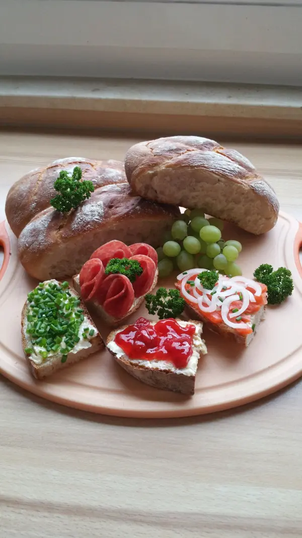 Vier knusprige Brötchen liegen auf einem Teller, umgeben von bunten belegten Broten und Trauben.