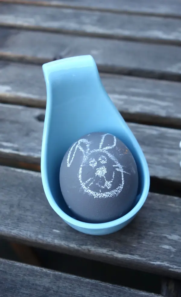 Ein schwarz bemaltes Osterei liegt in einem blauen Halter und zeigt mit Kreide ein Häschen. Es ist auf einer Holzoberfläche platziert.