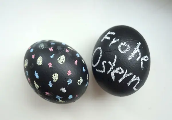 Zwei Ostereier aus schwarzem Tafellack, eines mit bunten Mustern, das andere mit „Frohe Ostern“ in Kreide beschriftet.