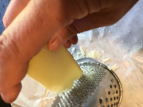 Eine Hand reibt einen Käseblock über eine feine Reibe, während ein Bärlauchpesto in einer Schüssel zubereitet wird.