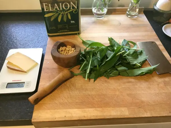 Auf einem Holzbrett liegt frischer Bärlauch neben einer Schüssel mit Nüssen, einer Käsescheibe und Olivenöl.