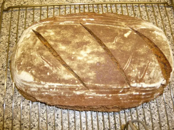 Ein frisch gebackenes Sauerteig-Roggenmischbrot mit einer goldbraunen Kruste liegt auf einem Kuchengitter in einer hellen Küche.