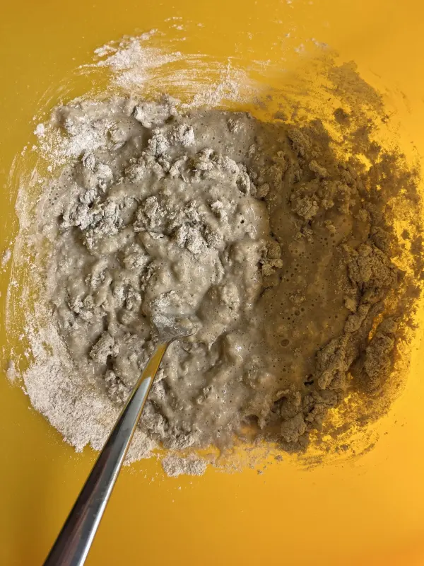 Grau-brauer Teig wird in einer gelben Schüssel mit einem Löffel gerührt, um Sauerteig-Roggenmischbrot vorzubereiten.