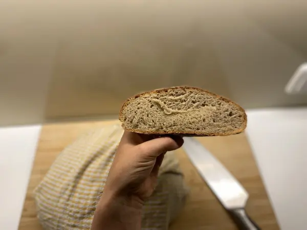 Ein handgeschnittenes Sauerteig-Roggenmischbrot wird vor einem Holzbrett gehalten, mit einem Messer daneben.