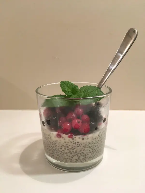 Ein Glas mit Chia-Samen-Pudding, garniert mit frischen Beeren und Minze, steht auf einem weißen Tisch. Ein Löffel ist darin.
