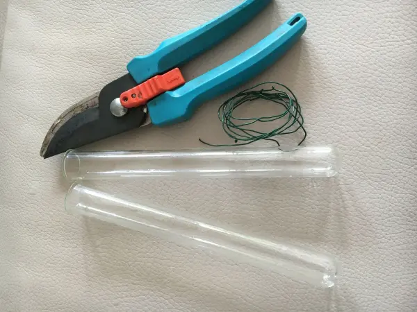 Zwei Glasröhren liegen zusammen mit einer Schere und Draht auf einem hellen Untergrund, bereit für kreativen Fensterschmuck.
