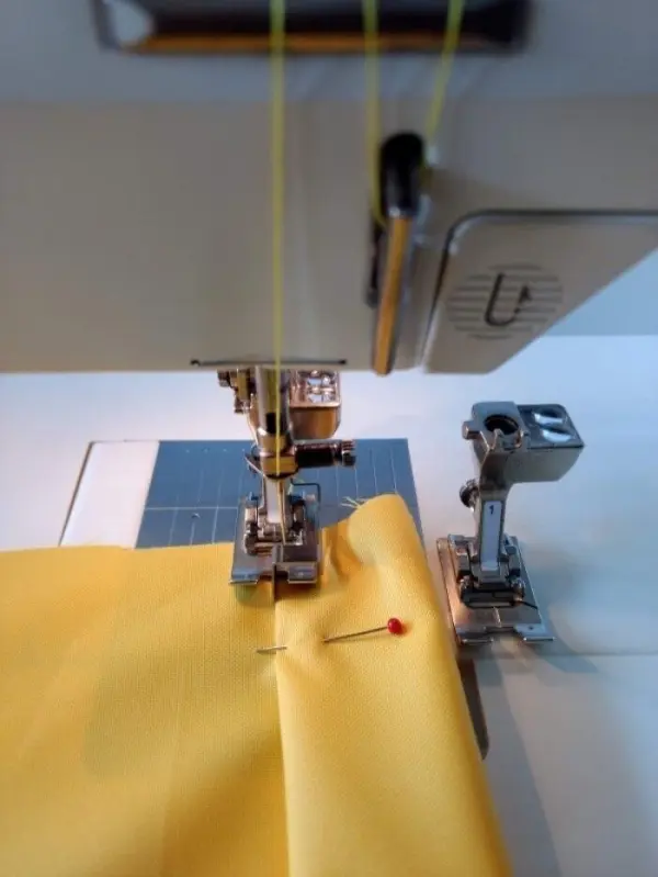 Eine gelbe Stoffbahn wird an einer Nähmaschine genäht, während sie mit einer Stecknadel fixiert ist, auf einem Tisch mit Schnittmatte.