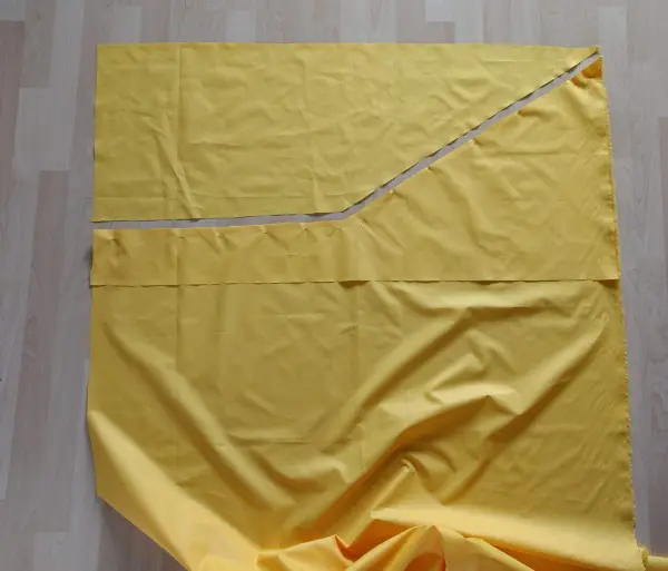 Ein gelbes Stoffstück liegt auf einem Boden. Eine Linie zeigt die Schnittform für Sonnen- und Windschutz.
