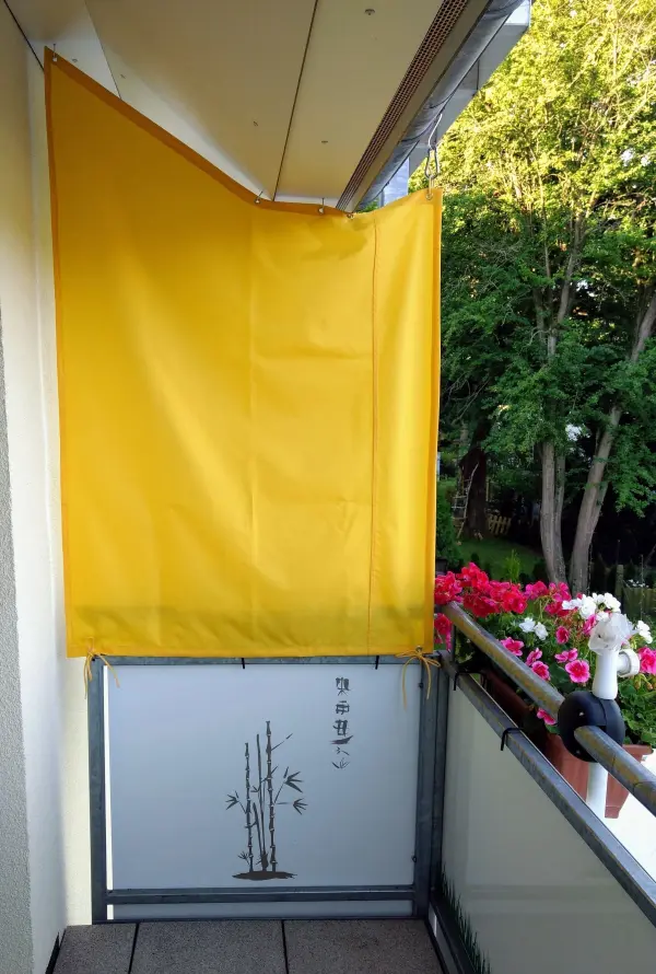 Ein gelbes Sonnenschutz-Paneel hängt auf einem Balkon, um Schatten zu spenden, umgeben von bunten Blumen.