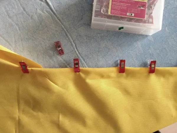 Ein gelbes Stoffstück wird mit roten Clips am Rand fixiert, während es auf einem blauen Untergrund zum Nähen vorbereitet wird.