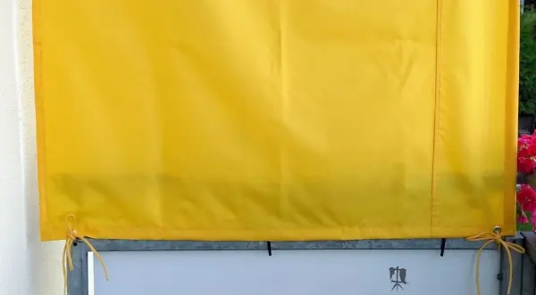 Ein gelbes Sonnenschutzvorhang hängt an einem Fenster und bietet Schatten in einer blühenden Umgebung.