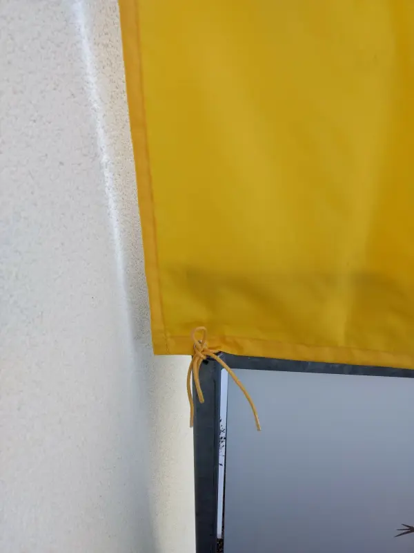 Ein gelbes Sonnenschutzprodukt hängt an einem Rahmen, während es an einer Wand befestigt ist und Schatten spendet.