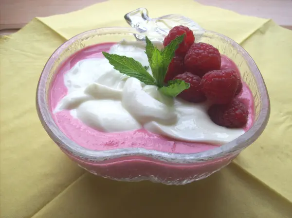 Ein pinkes Quark-Frischkäse-Dessert in einer Schale, garniert mit Himbeeren und Minze, auf einem gelben Untergrund.