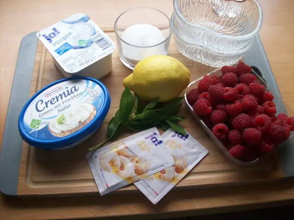 Ein Quark-Frischkäse-Dessert wird mit frischen Himbeeren, Zitronen und weiteren Zutaten auf einem Holzbrett vorbereitet.