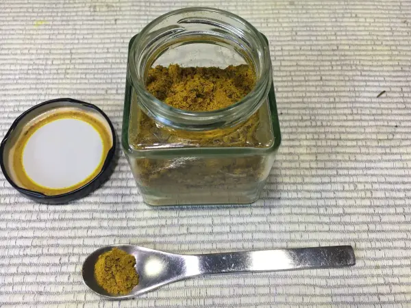 Ein Glasbehälter mit Kurkuma steht offen auf einer Tischoberfläche, während ein Löffel Kurkuma daneben platziert ist.