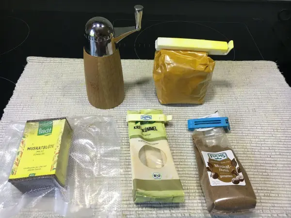 Ein Glasbehälter mit Kurkuma steht neben einem Öler, während verschiedene Gewürzverpackungen auf einer weißen Unterlage angeordnet sind.