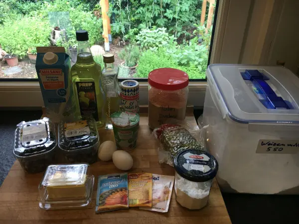 Auf einem Holzbrett liegen Zutaten für schwedische Blaubeer-Tartelettes: Eier, Beeren, Mehl und Gewürze sind sichtbar, im Hintergrund ein Garten.