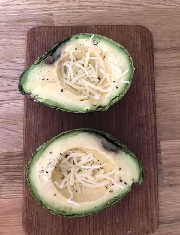 Eine auf einem Holzbrett liegende, halbierte Avocado, die mit Käse bestreut ist und auf das Backen vorbereitet wird.