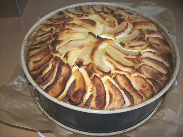 Ein Käsekuchen mit goldbraunen Rum-Apfelschnitzen, frisch gebacken, steht auf Backpapier in einer runden Springform.