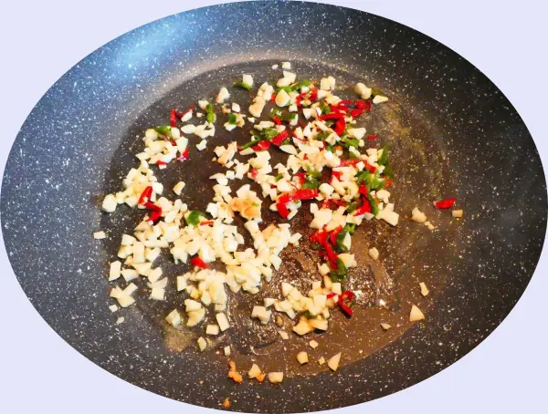 In einer schwarzen Pfanne brutzeln gehackter Knoblauch und rote Chilischoten in Öl; scharfe Aromen entfalten sich.