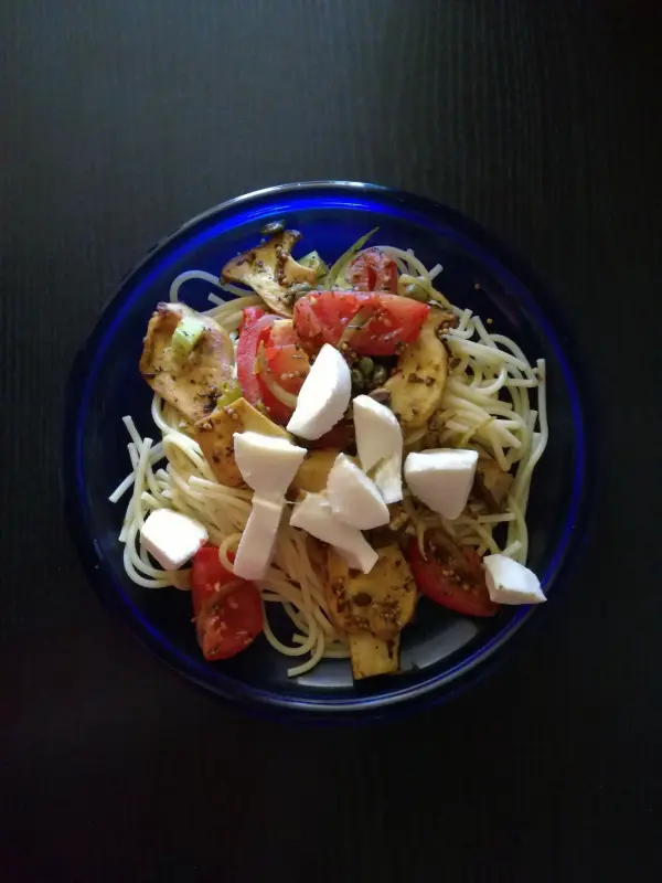 Eine bunte Schüssel Spaghetti mit Kräuterseitlingen, frischen Tomaten und Mozzarella auf einem dunklen Tisch.