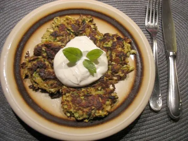 Knusprige Zucchini-Rösti liegen auf einem Teller, garniert mit einem Klecks Sahne und frischen Minzblättern.