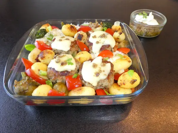 Käseüberbackene Pesto-Frikadellen liegen auf einem bunten Gemüsebett aus Kartoffeln, Tomaten und Brokkoli in einer Glasschale.