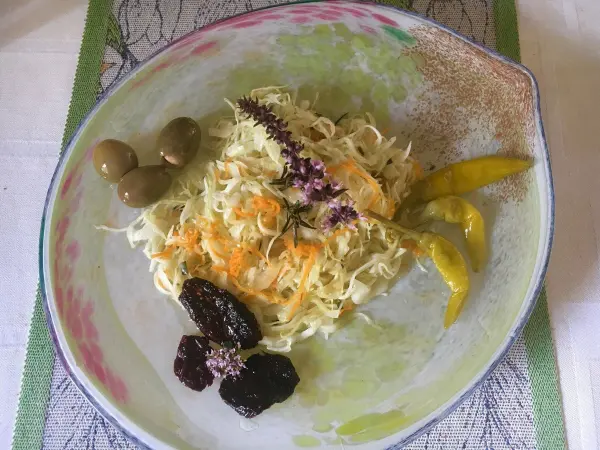 Ein frisch zubereiteter Krautsalat liegt auf einem Teller, umgeben von grünen Oliven und gelben Paprikastreifen.
