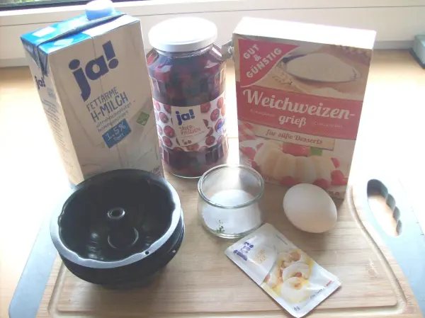 Grießflammeri-Zutaten wie Grieß, Kirschkompott und Zucker liegen auf einem Holzbrett in einer hellen Küche.