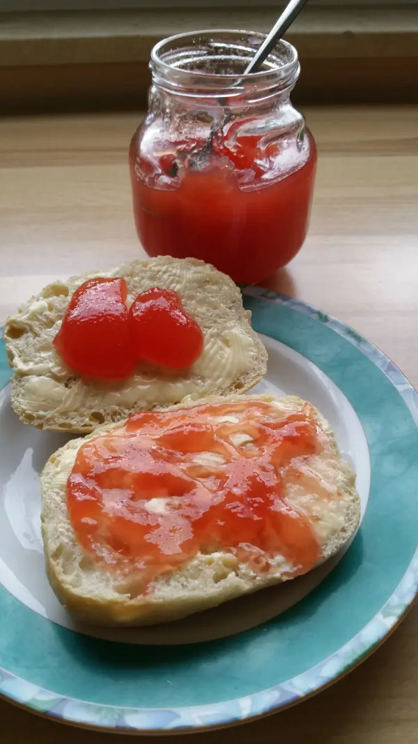 Eine Scheibe Brot ist mit fruchtiger Wassermelonenmarmelade bestrichen, während ein Glas mit Marmelade im Hintergrund steht.