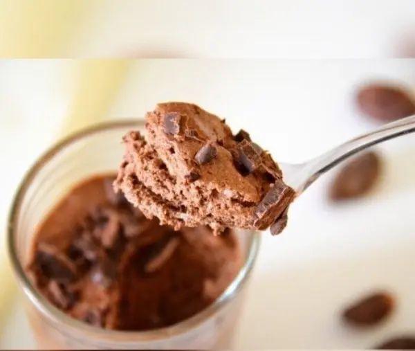 Eine Löffelportion Mousse au Chocolat hebt sich über einem Glas, umgeben von Schokoladenstückchen auf einem hellen Hintergrund.