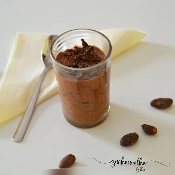 Eine Glasportion Mousse au Chocolat steht auf einem hellen Untergrund, umgeben von Schokoladenstückchen und einem Löffel.
