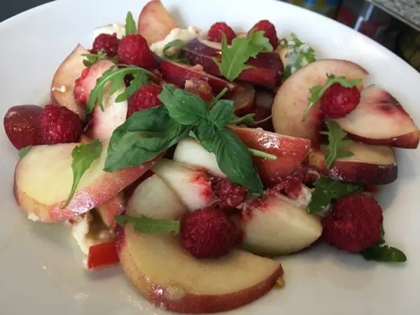 Ein bunter Salat aus Nektarinen, frischen Himbeeren und Mozzarella wird mit Basilikum garniert, präsentiert auf einem weißen Teller.