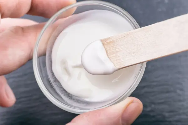 Eine Hand hält einen Holzspatel, der eine dicke, weiße Paste aus einem Glasbehälter hebt, um Fettspritzer an der Wand zu entfernen.
