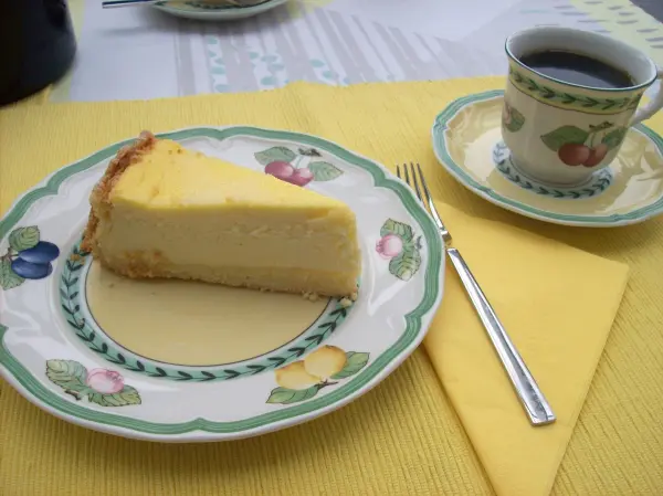 Ein Stück perfekter Käsekuchen liegt auf einem bunten Teller neben einer Tasse Kaffee auf einem gelben Tischset.
