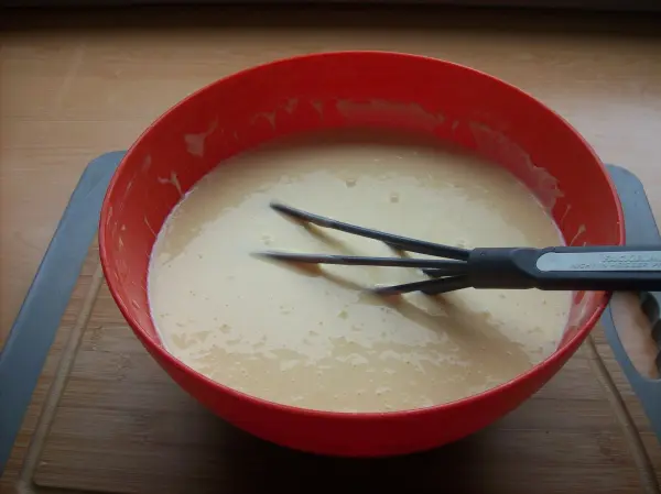 In einer roten Schüssel wird ein cremiger Käsekuchenmix mit einem Schneebesen angerührt, auf einem Holzbrett platziert.