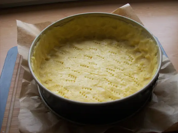 Ein unbebackener Boden für einen Käsekuchen liegt in einer runden Springform, umgeben von Backpapier auf einer Holzplatte.