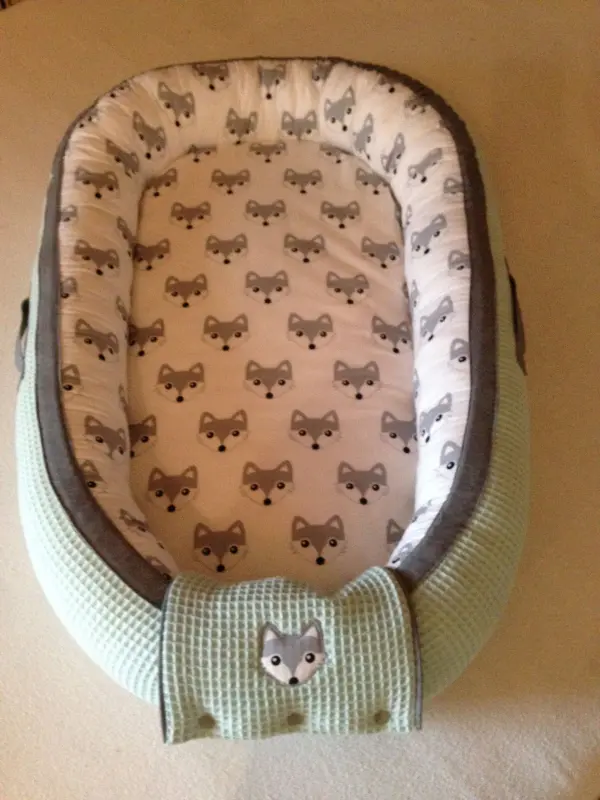Ein Babynest aus grauem und mintgrünem Stoff mit niedlichem Fuchs-Muster liegt auf einem hellen Untergrund.