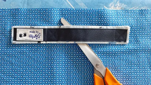 Eine Schere schneidet ein schwarzes Label auf einem blauen Stoff mit Punkten, während ein Babynest genäht wird.