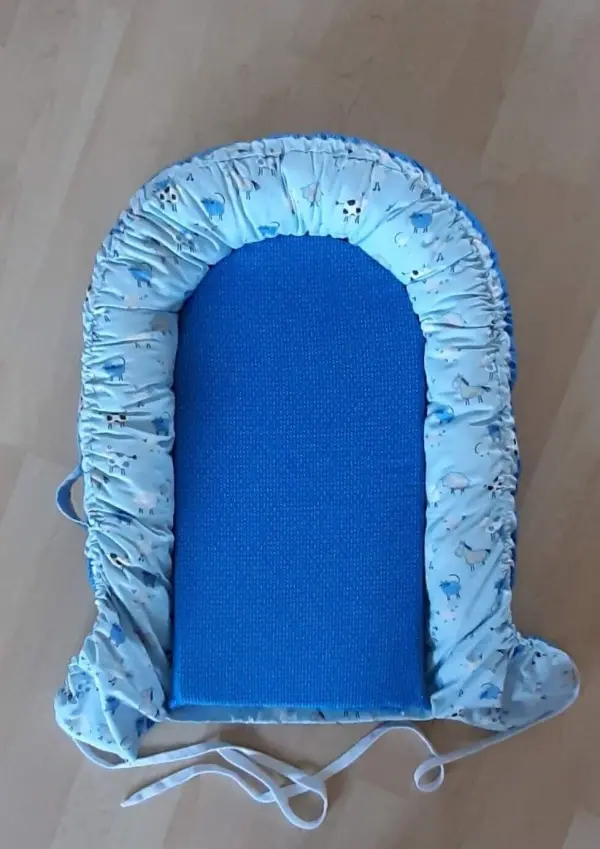 Ein Babynest mit blauer Matratze und einem gepolsterten Rand aus bedrucktem Stoff liegt auf einem hellen Boden.